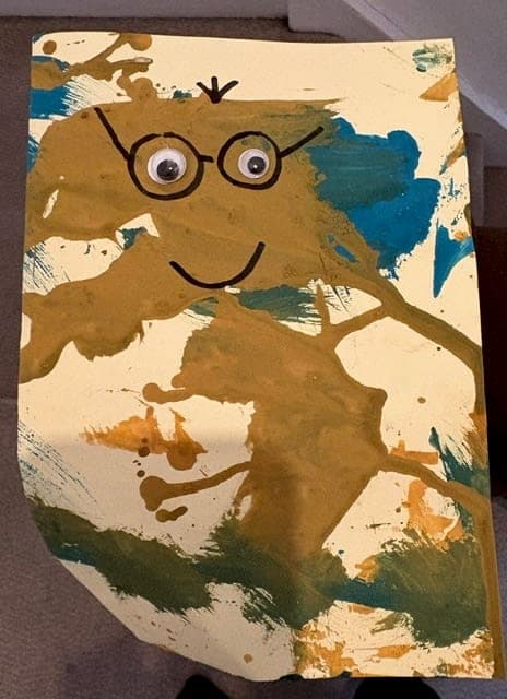 A phone photo of a child's painting lying on a carpet: a large brown paint blob on cream card, with googly eyes glued on and round glasses and a smile drawn over it in black marker.