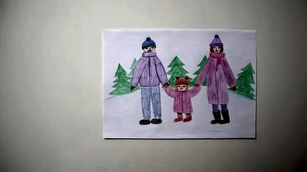 A phone photo of a child's felt-tip drawing of a family of three in winter coats and bobble hats, holding hands between fir trees, on white paper against a dark background.